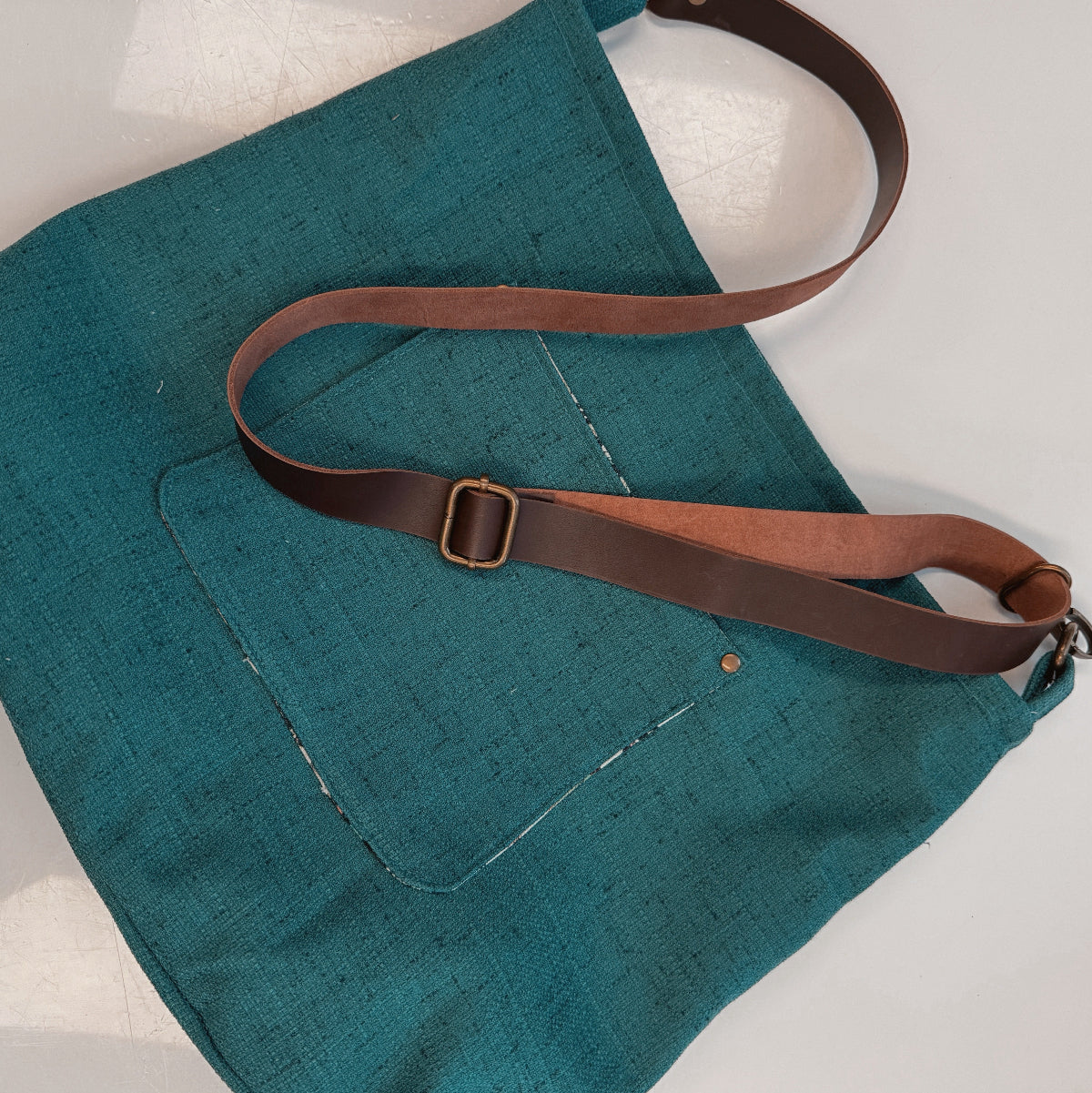 A teal woven fabric shoulder bag with a brown leather strap and brass buckle, laid flat on a white surface.