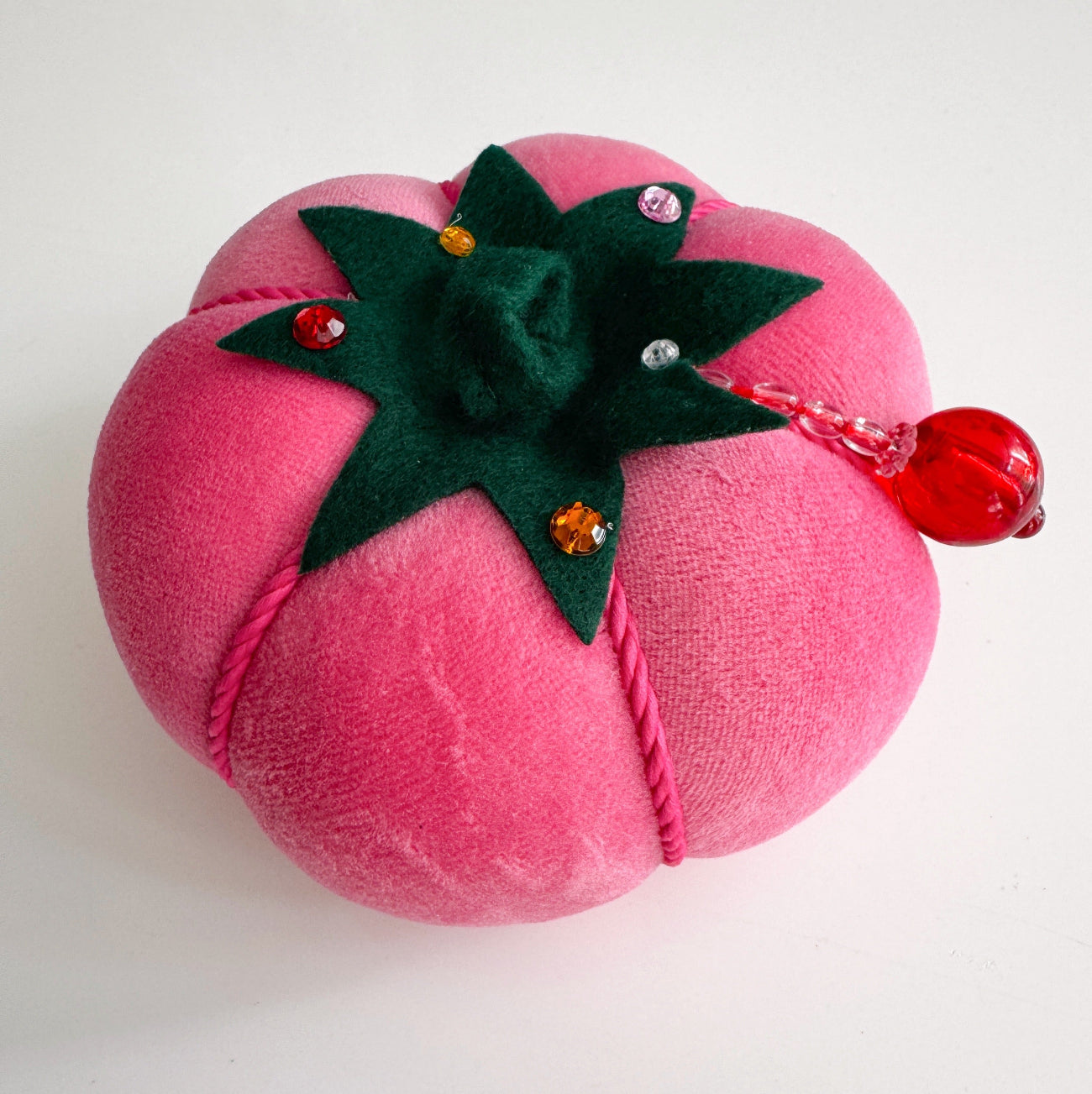 A person’s hand holding a round, bright pink velvet tomato pincushion adorned with a dark green stem, decorative colored crystals, and a small red beaded accent.