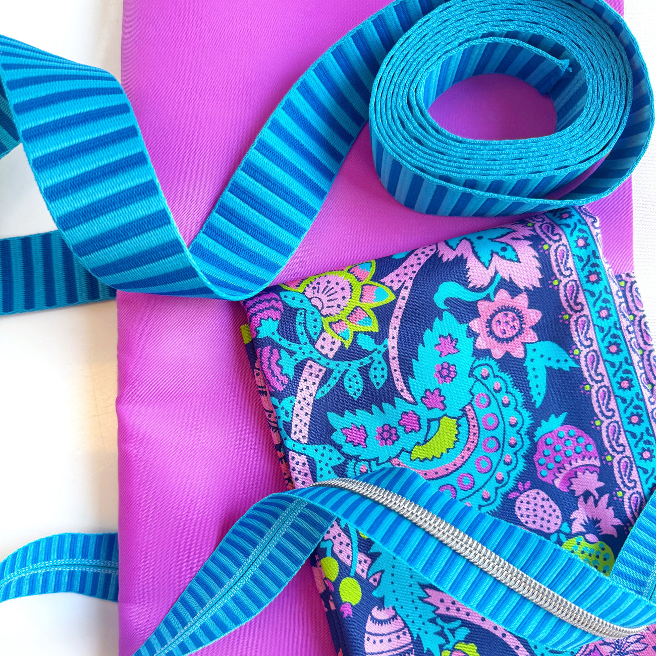 SewHungryHippie MATCHUP 1 sewing bundle showcasing a folded yard of JPag fabric with a busy bohemian floral and paisley pattern in navy blue, bright pink, teal, and lime green. The fabric rests on a large piece of solid bright magenta bonded nylon, accompanied by coiled lengths of blue striped webbing and a matching blue striped zipper tape with silver teeth.