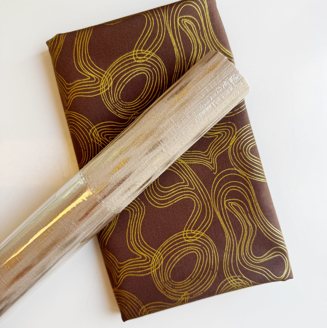 SewHungryHippie MATCHUP 10 craft bundle showing a roll of metallic Gold Bands vinyl laid diagonally over a folded yard of dark brown fabric. The fabric features an elegant, swirling pattern of thin, concentric gold accent lines resembling topographic maps or abstract waves.