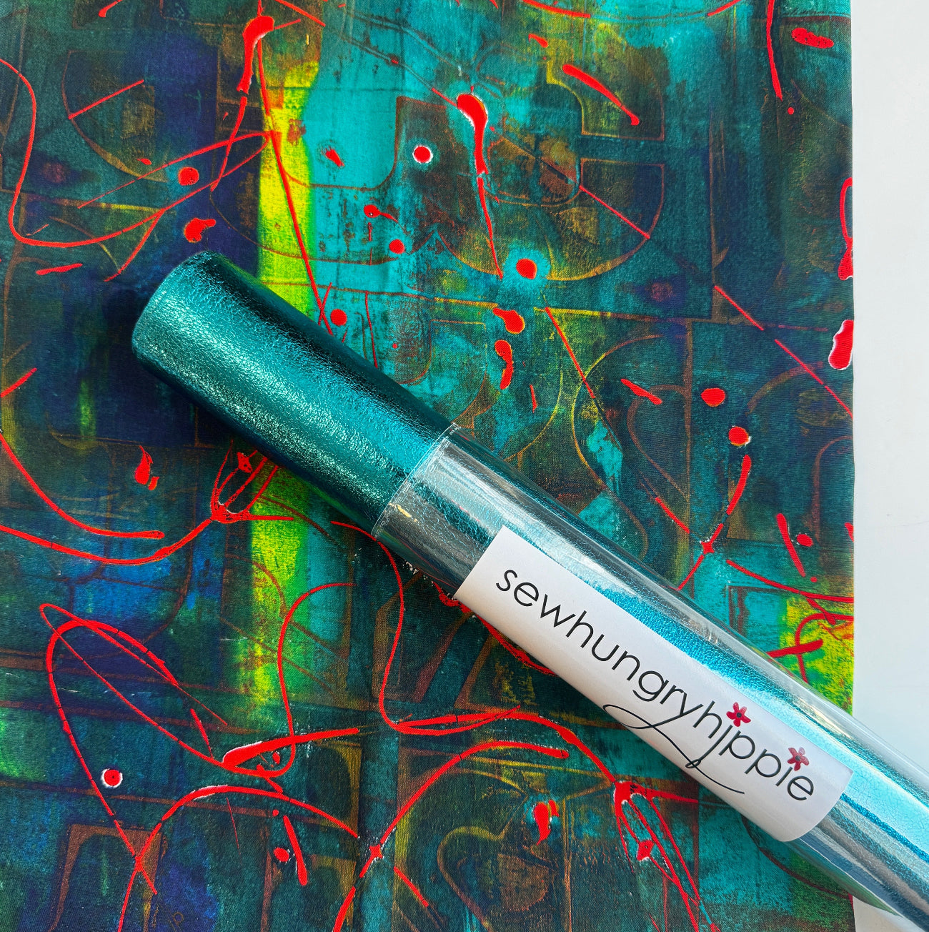 SewHungryHippie MATCHUP 2 bundle featuring a rolled tube of metallic teal soft vinyl with a white brand label, resting on top of a flat yard of Graphic Warehouse fabric. The fabric displays an abstract design with a dark teal, blue, and lime green textured background accented by bright red splatters and scribbled lines.
