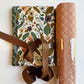 SewHungryHippie MATCHUP 4 craft bundle containing a roll of peach-colored quilted vinyl next to two folded yards of floral fabric featuring an earth-toned pattern of muted oranges, purples, and olive green leaves on a cream background. A coiled brown leather strap loops across the front of the fabric.