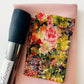 SewHungryHippie MATCHUP 5 sewing bundle featuring a folded piece of light pink shimmer fabric as the base. On top rests a smaller cut of Tim Holtz fabric with an impressionistic, multi-colored floral pattern in pink, yellow, red, and green. A roll of textured Navy Moonglow vinyl with a brand label lies vertically along the left side.