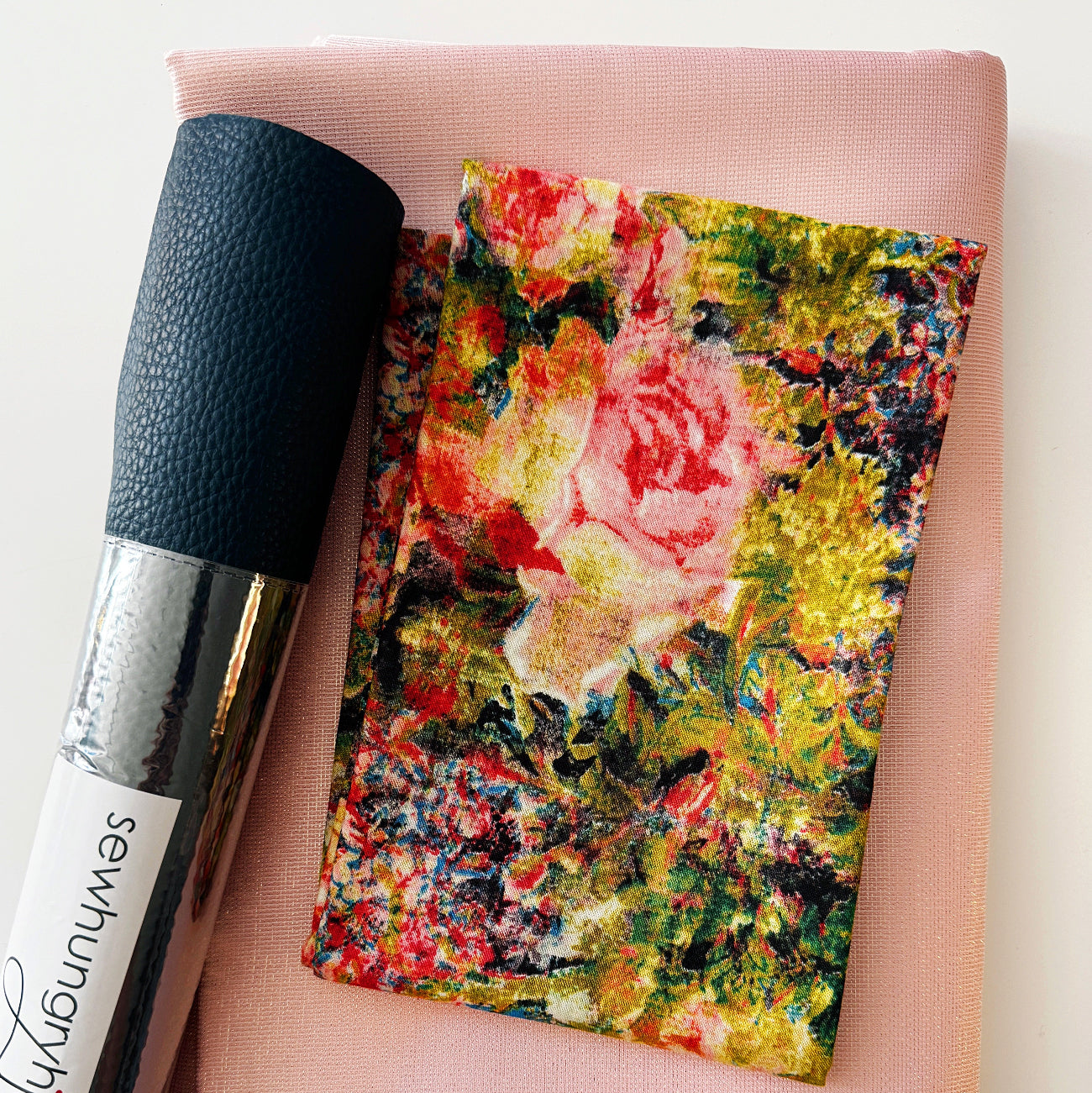 SewHungryHippie MATCHUP 5 sewing bundle featuring a folded piece of light pink shimmer fabric as the base. On top rests a smaller cut of Tim Holtz fabric with an impressionistic, multi-colored floral pattern in pink, yellow, red, and green. A roll of textured Navy Moonglow vinyl with a brand label lies vertically along the left side.
