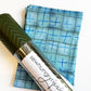 SewHungryHippie MATCHUP 7 craft bundle featuring a roll of olive green quilted chevron vinyl with a white brand label. The roll is positioned diagonally over a folded yard of light blue and teal fabric decorated with a grid pattern and hand-drawn blue plus signs.