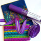 SewHungryHippie MATCHUP 8 sewing bundle featuring two different cuts of Sally K fabric: a large royal blue piece adorned with a vibrant, colorful whimsical garden border print, and a folded piece with horizontal rows of bright pink, purple, yellow, teal, and green leaf vines. A roll of textured Moonglow Purple vinyl with a brand label rests diagonally across the top, accompanied by a coiled length of purple striped zipper tape with silver teeth.