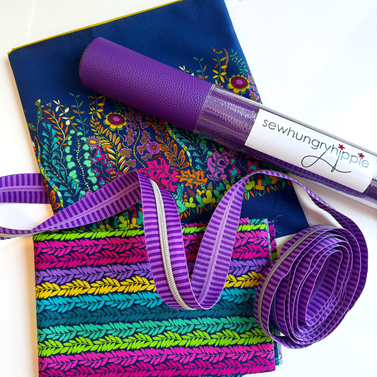 SewHungryHippie MATCHUP 8 sewing bundle featuring two different cuts of Sally K fabric: a large royal blue piece adorned with a vibrant, colorful whimsical garden border print, and a folded piece with horizontal rows of bright pink, purple, yellow, teal, and green leaf vines. A roll of textured Moonglow Purple vinyl with a brand label rests diagonally across the top, accompanied by a coiled length of purple striped zipper tape with silver teeth.