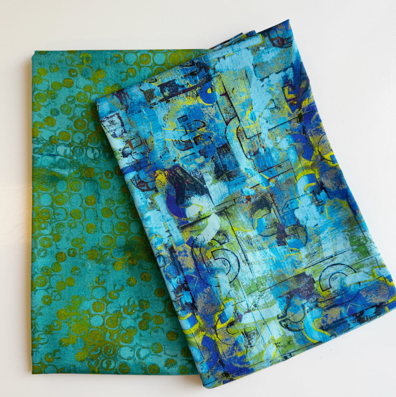 Two folded yards of SewHungryHippie MATCHUP 9 fabric coordinates. The bottom fabric features an abstract green-and-teal bubble pattern, while the top fabric displays a vibrant, textured abstract design in shades of blue, teal, purple, and lime green.