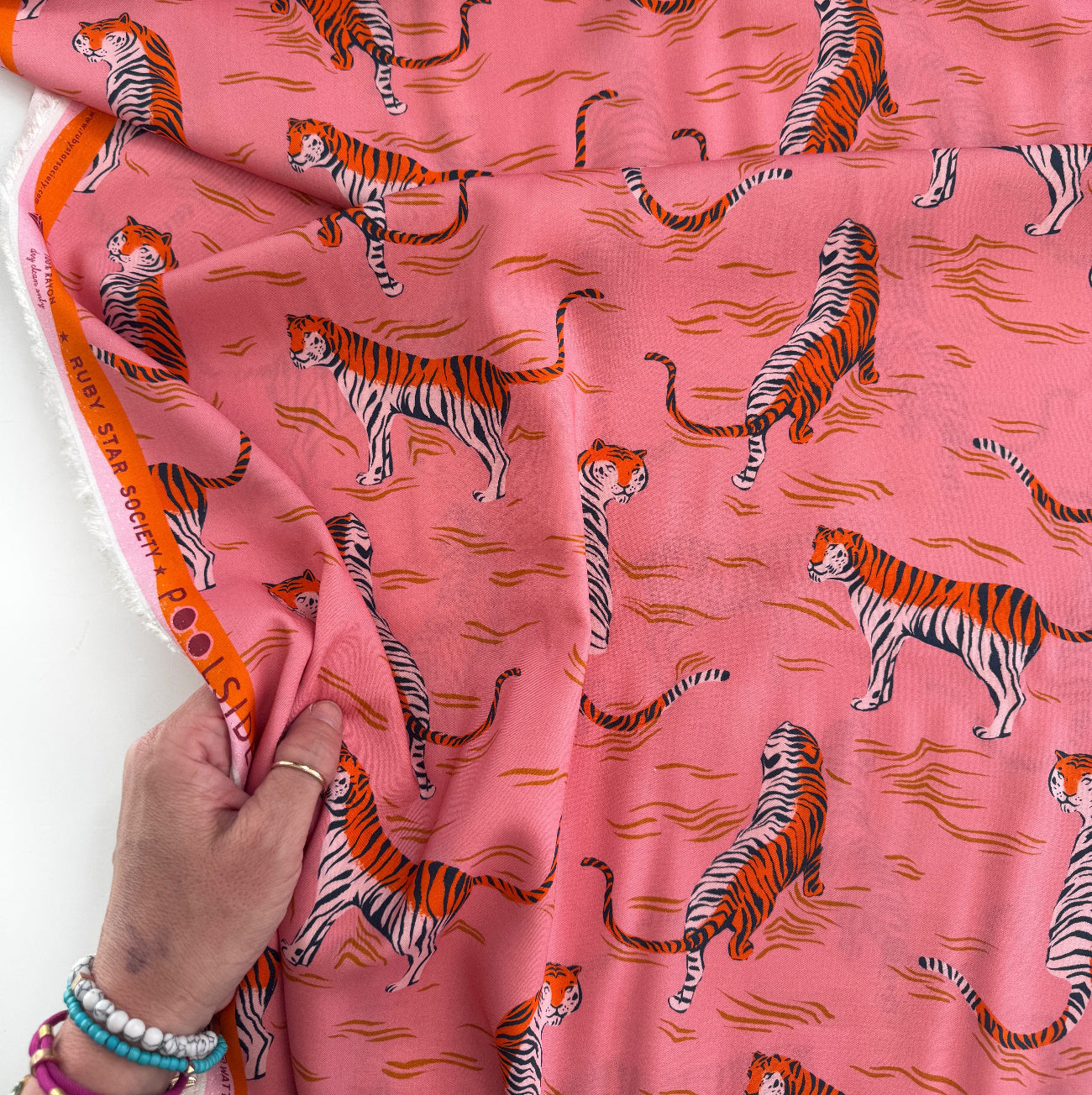 A person’s hand holds a draped yard of Ruby Star Society rayon fabric, displaying a repeating pink, orange, and black tiger pattern with a branded orange selvage edge.