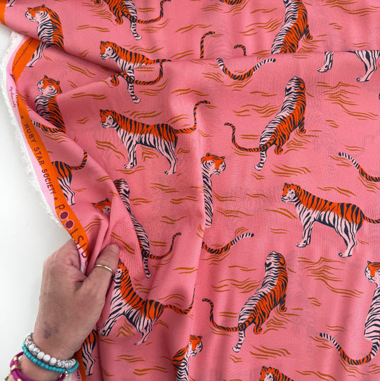 A person’s hand holds a draped yard of Ruby Star Society rayon fabric, displaying a repeating pink, orange, and black tiger pattern with a branded orange selvage edge.