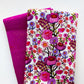 Two folded pieces of fabric on a white background, one with a white base covered in a dense pink, purple, and green floral print, and the other in a solid magenta-pink with subtle gold speckles.