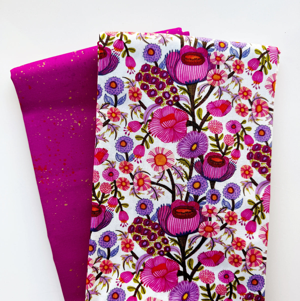 Two folded pieces of fabric on a white background, one with a white base covered in a dense pink, purple, and green floral print, and the other in a solid magenta-pink with subtle gold speckles.