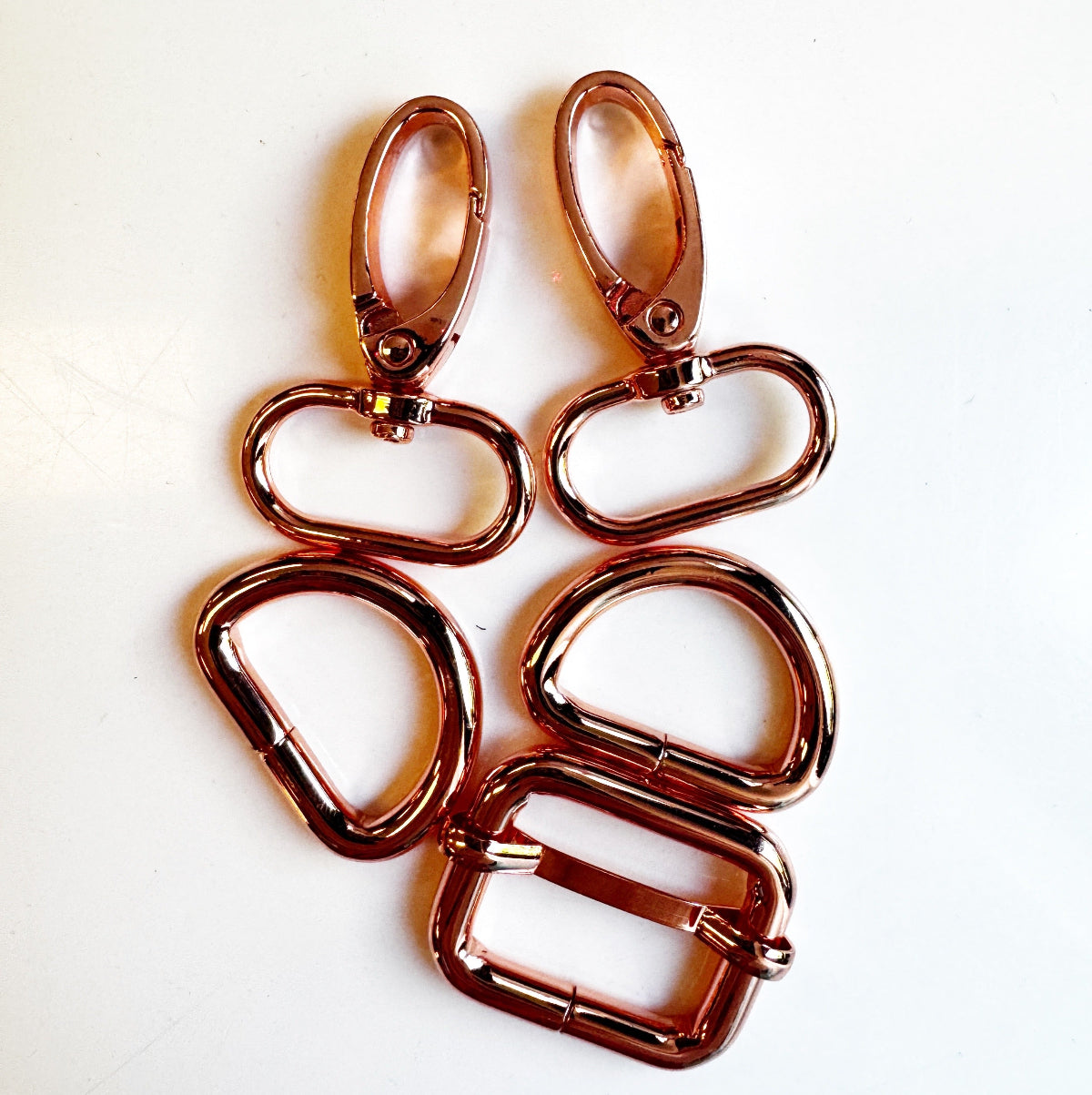 Four rose gold metal hardware components, including two lobster clasps and two D-rings, arranged on a plain white background.