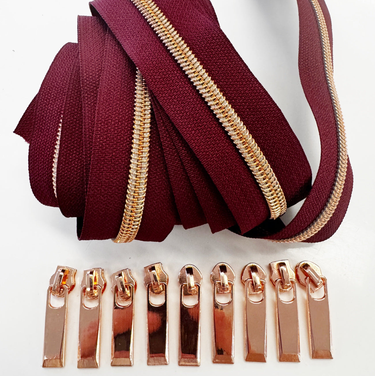 A set of burgundy fabric zipper tapes with gold-toned metal teeth, displayed above matching rose gold metal zipper pull tabs on a plain white background.