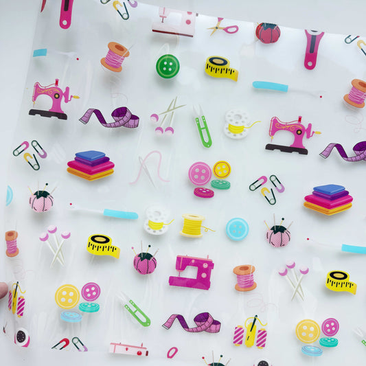 A Sewing Notions Clear Vinyl TPU by SewHungryhippie, featuring a transparent design printed with colorful sewing machines, spools of thread, buttons, and tape measures.