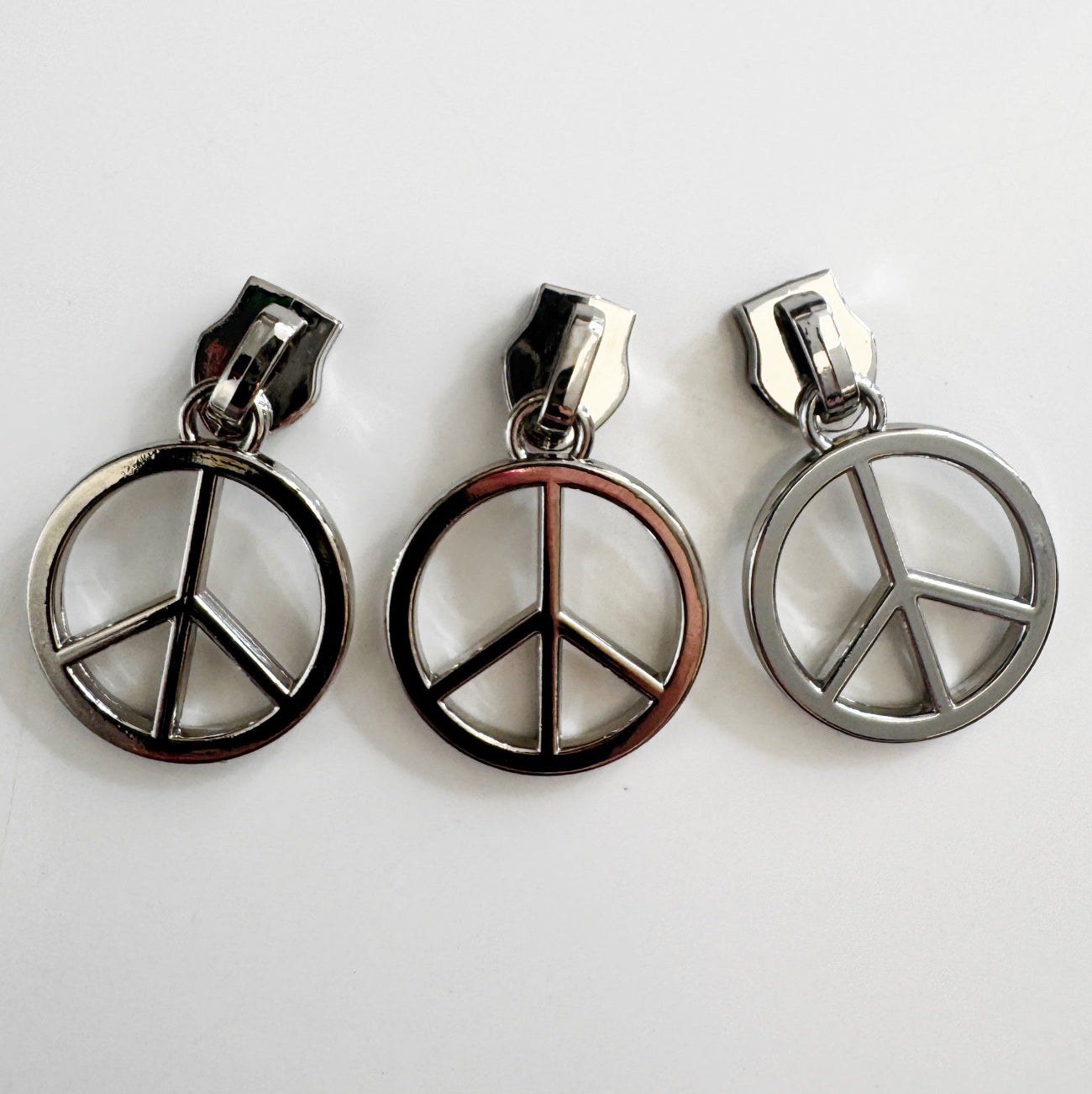 Three metallic silver-tone zipper pulls featuring a classic circular peace sign charm, aligned side-by-side against a plain white background.