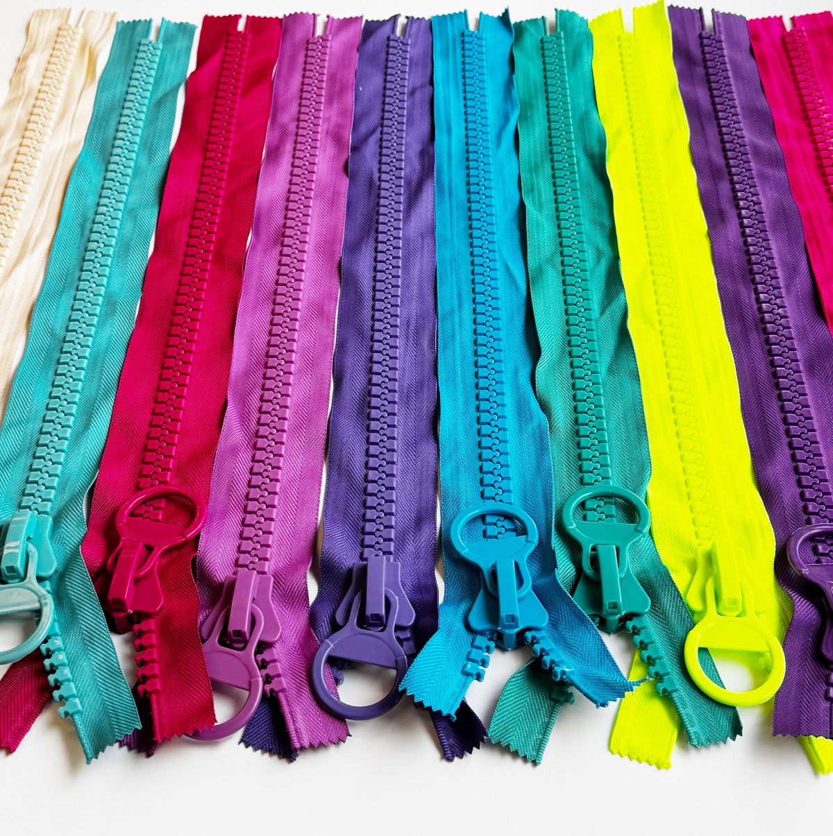 A set of colorful metal zipper teeth strips arranged vertically in a rainbow-like gradient on a white background, including cream, teal, pink, purple, blue, and neon yellow.
