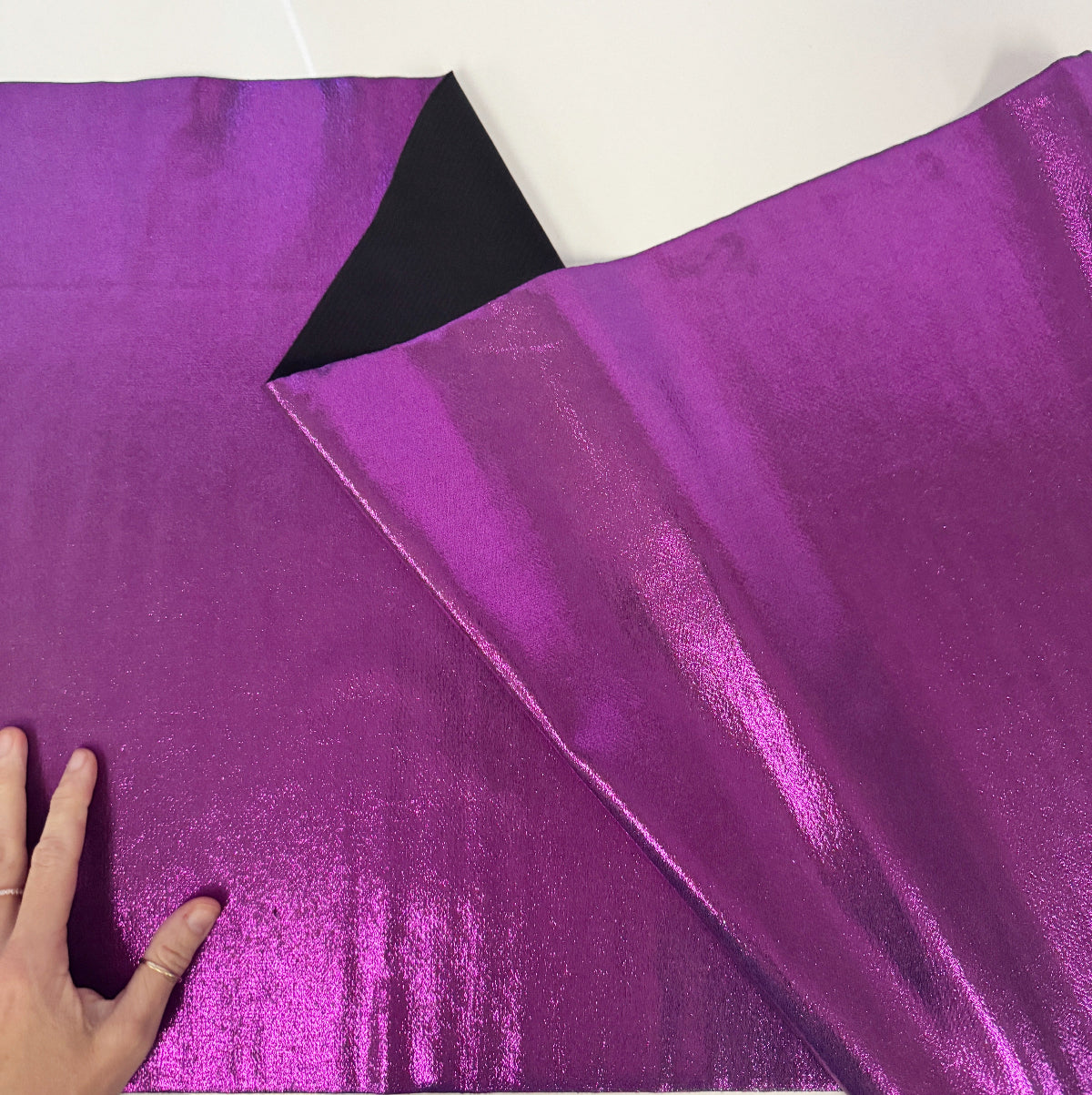A close-up of shiny metallic purple fabric, with one corner folded over to show the reverse side is black. A hand is visible holding the fabric at the lower left to show scale.