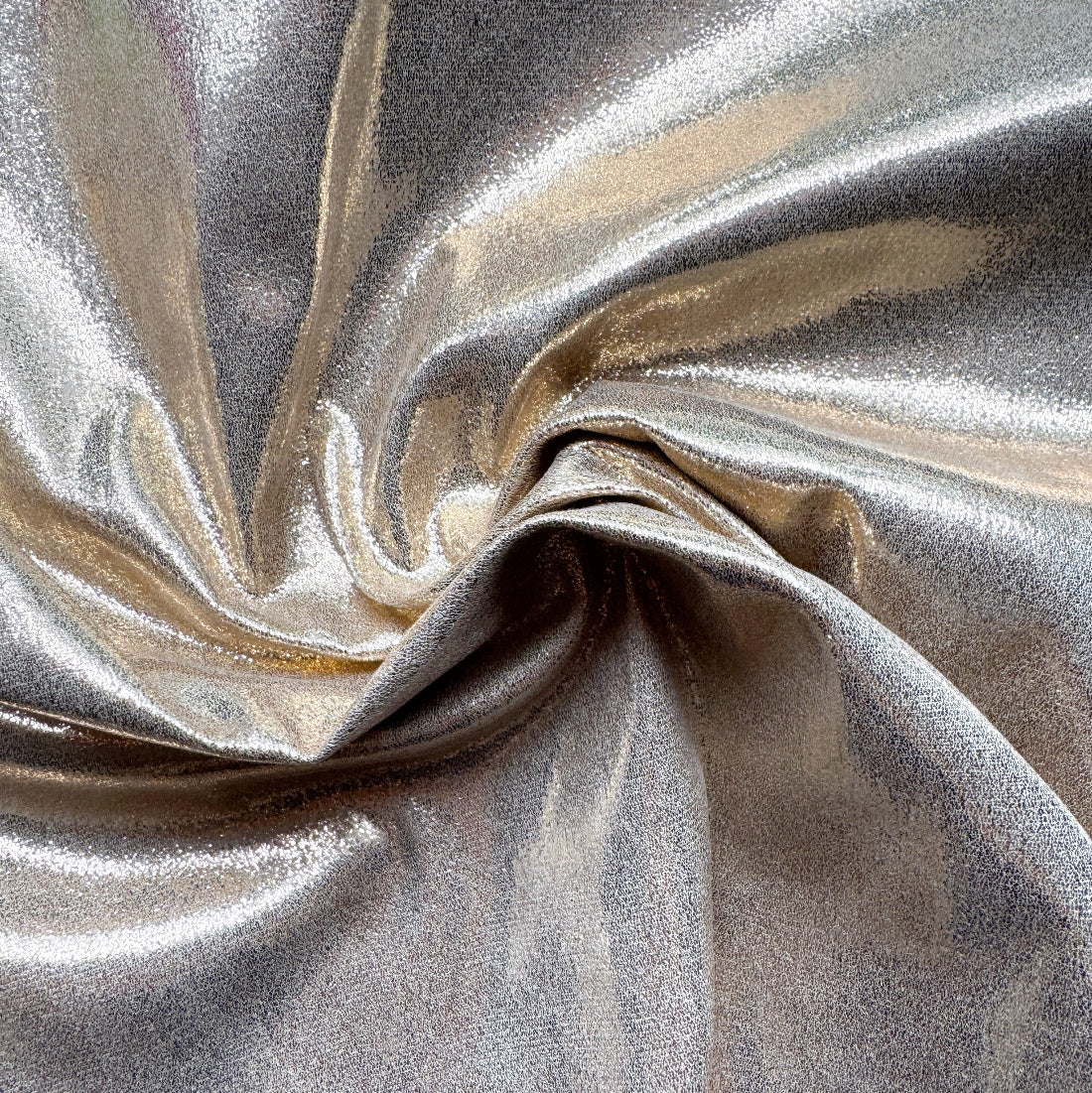 A close-up of shimmering metallic fabric with a silver and champagne-gold sheen, arranged in soft folds that highlight its reflective texture.