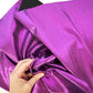 A close-up of shiny metallic purple fabric being held by a hand, with a small black fabric sample visible at the top edge.