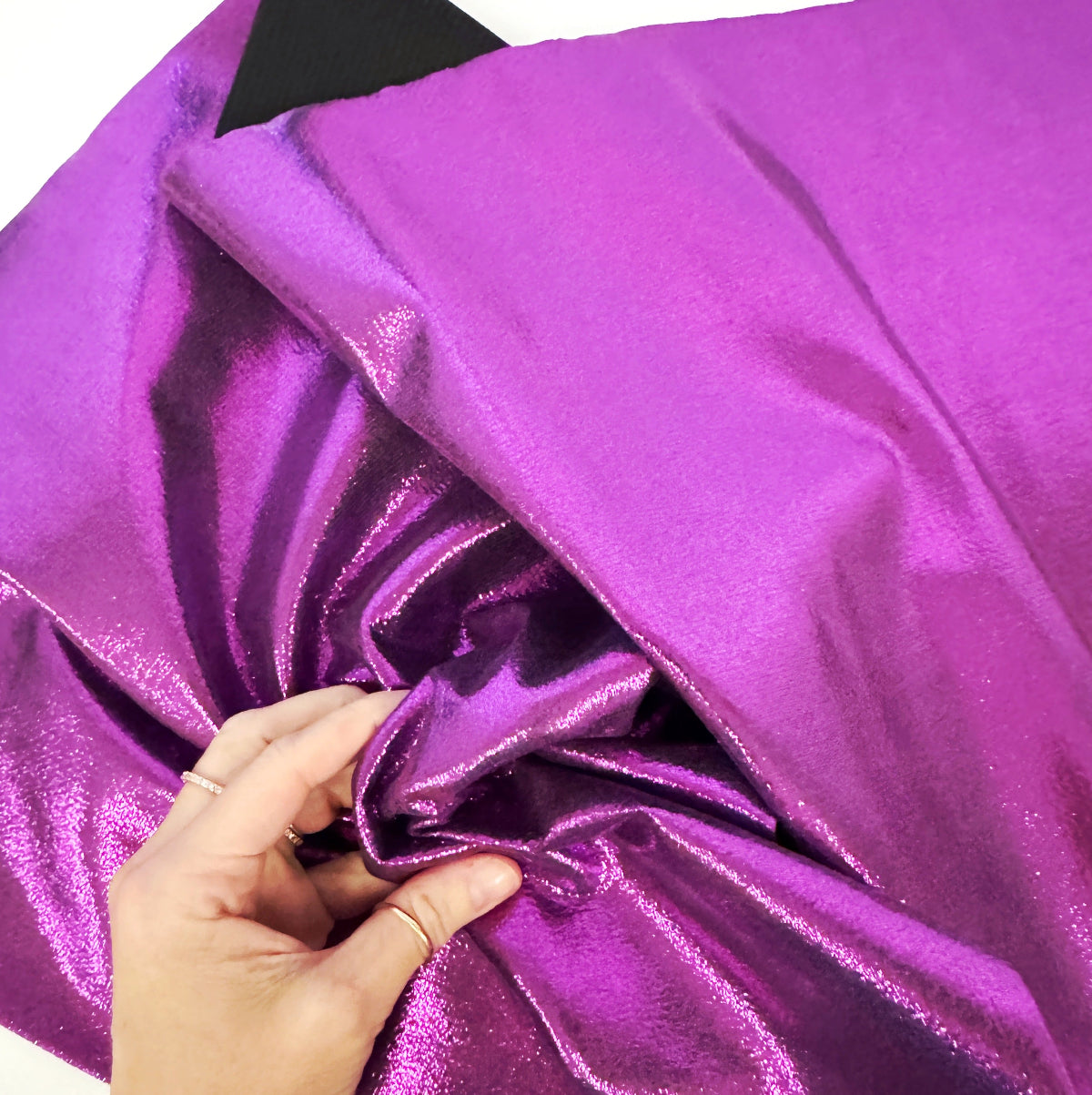 A close-up of shiny metallic purple fabric being held by a hand, with a small black fabric sample visible at the top edge.