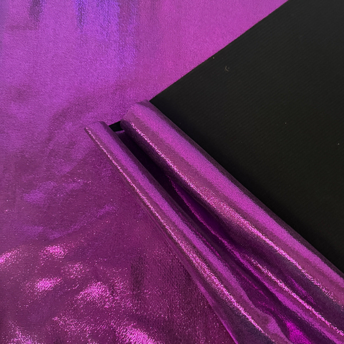 A close-up of shiny metallic purple fabric with a folded section revealing a black backing or second side, photographed against the same purple material.