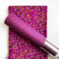 A purple glittery cosmetic tube, likely lip gloss or lip oil, resting diagonally on top of a folded fabric swatch with a matching magenta and yellow floral pattern.