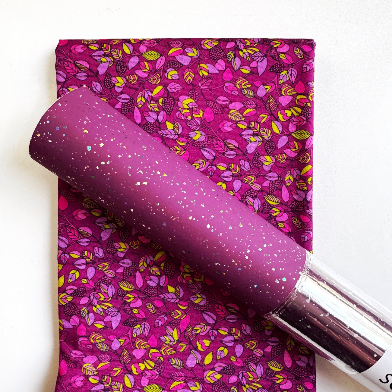 A purple glittery cosmetic tube, likely lip gloss or lip oil, resting diagonally on top of a folded fabric swatch with a matching magenta and yellow floral pattern.
