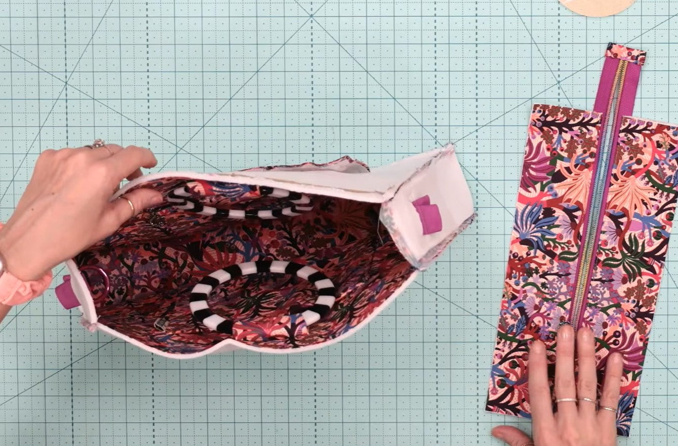 A top-down view of a person's hands holding open a floral zippered pouch to show the patterned lining, with the matching zippered pouch laid flat on a green cutting mat background.