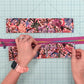 A person's hands are shown assembling a zipper case with a vibrant pink and multicolor floral pattern, placed on a green gridded cutting mat.