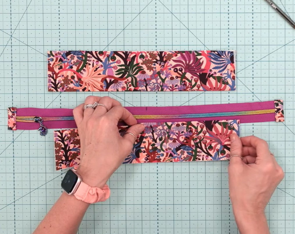 A person's hands are shown assembling a zipper case with a vibrant pink and multicolor floral pattern, placed on a green gridded cutting mat.