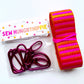 Bag-making supplies from Sew Hungry Hippie, including a hardware pack with fuchsia/magenta swivel clips and D-rings, paired with a roll of striped pink and orange webbing.