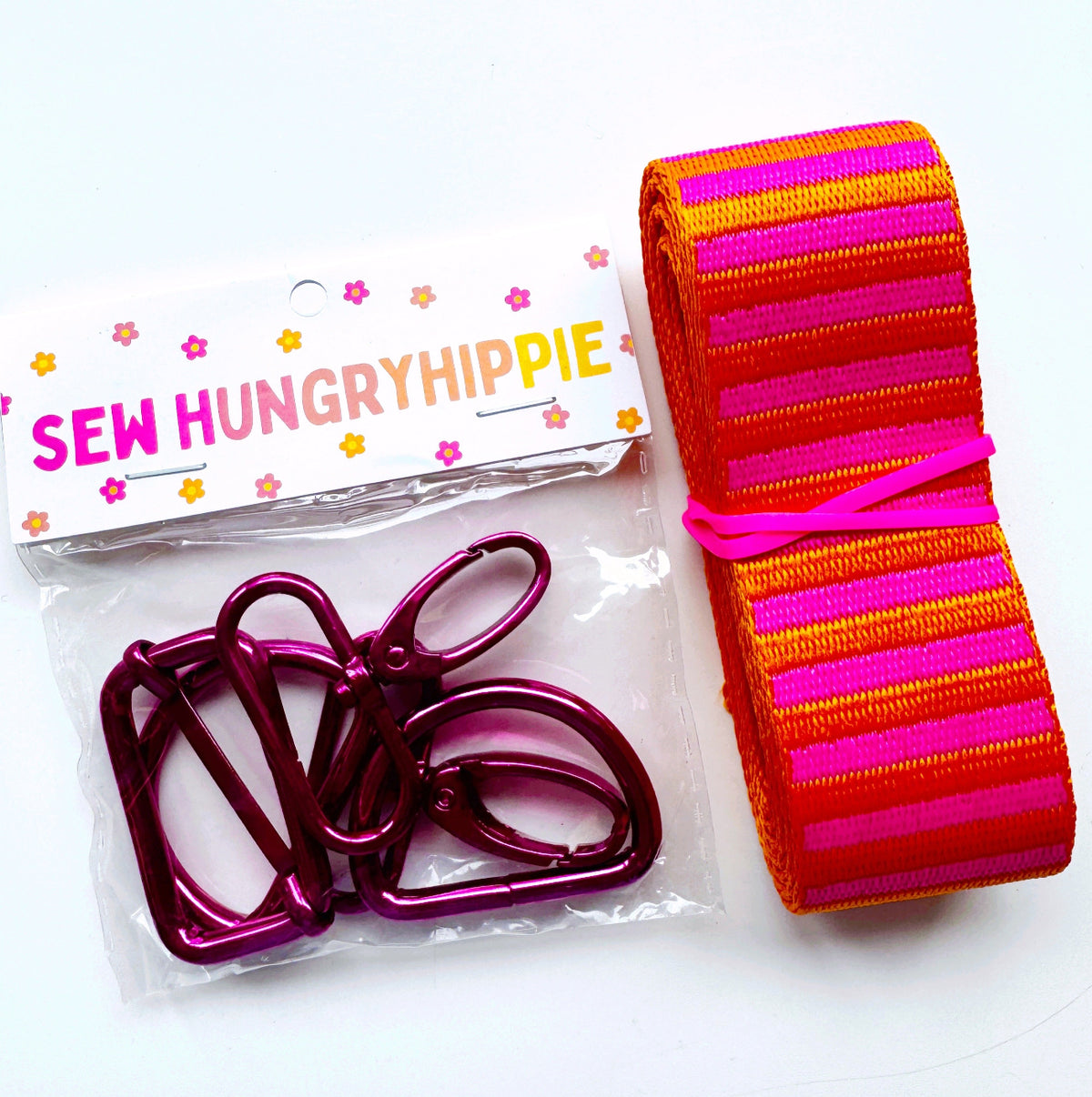 Bag-making supplies from Sew Hungry Hippie, including a hardware pack with fuchsia/magenta swivel clips and D-rings, paired with a roll of striped pink and orange webbing.