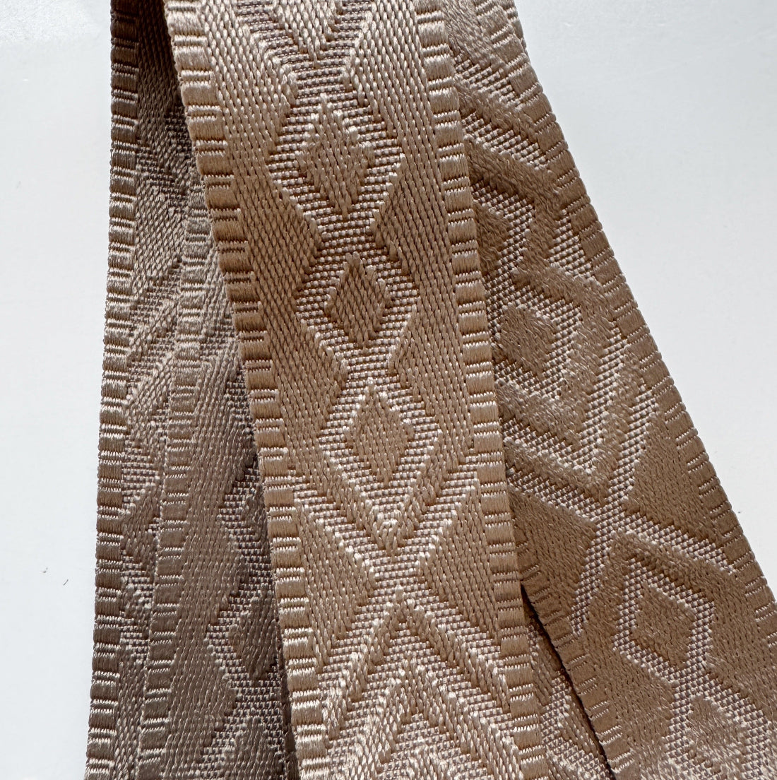 A close-up of a woven fabric strap or trim in a warm taupe-beige color, featuring a repeating geometric diamond pattern.