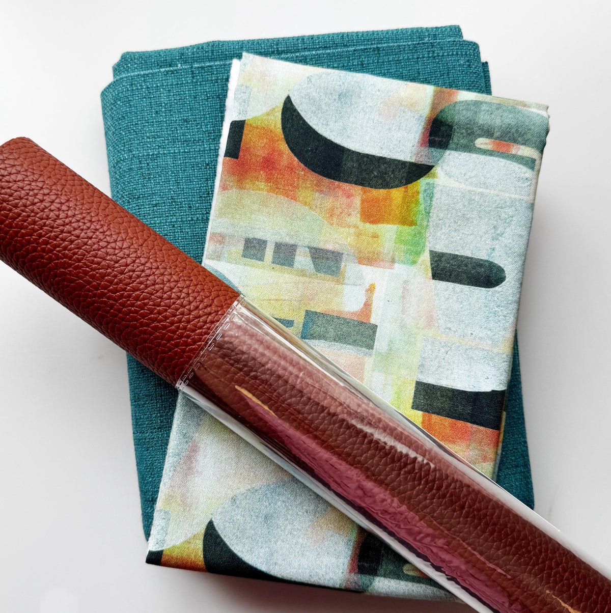 Teal fabric, abstract patterned fabric, and burgundy textured faux leather arranged together for a sewing or bag-making project.