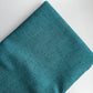 A close-up of folded teal woven fabric with a subtle textured weave, shown on a plain white background.