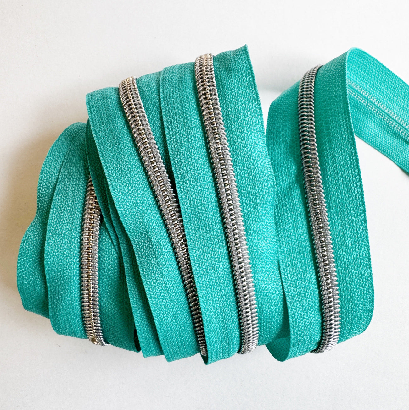 A close-up view of bright teal nylon zipper tape neatly layered and wrapped flatly to showcase several vertical, parallel rows of shiny silver metallic coil teeth against a plain white background.