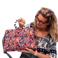 A woman with glasses and blonde hair holds up a colorful floral patterned duffel bag against a solid white background.