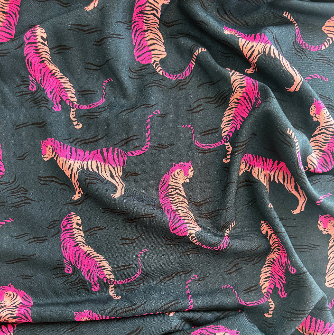 A close-up view of Rayon fabric featuring a dark teal background with a repeating pattern of stylized tigers in vibrant shades of pink and orange, accompanied by small black wavy lines.