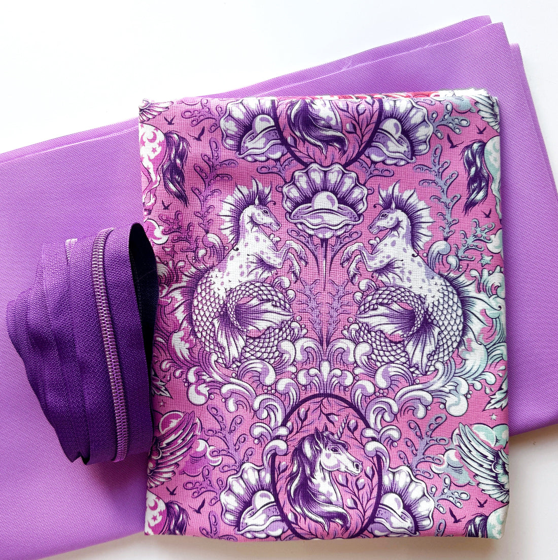A folded purple and pink patterned fabric item with a detailed floral and bird design, placed on top of solid purple fabric sheets. A matching purple zipper pull is visible on the left side.