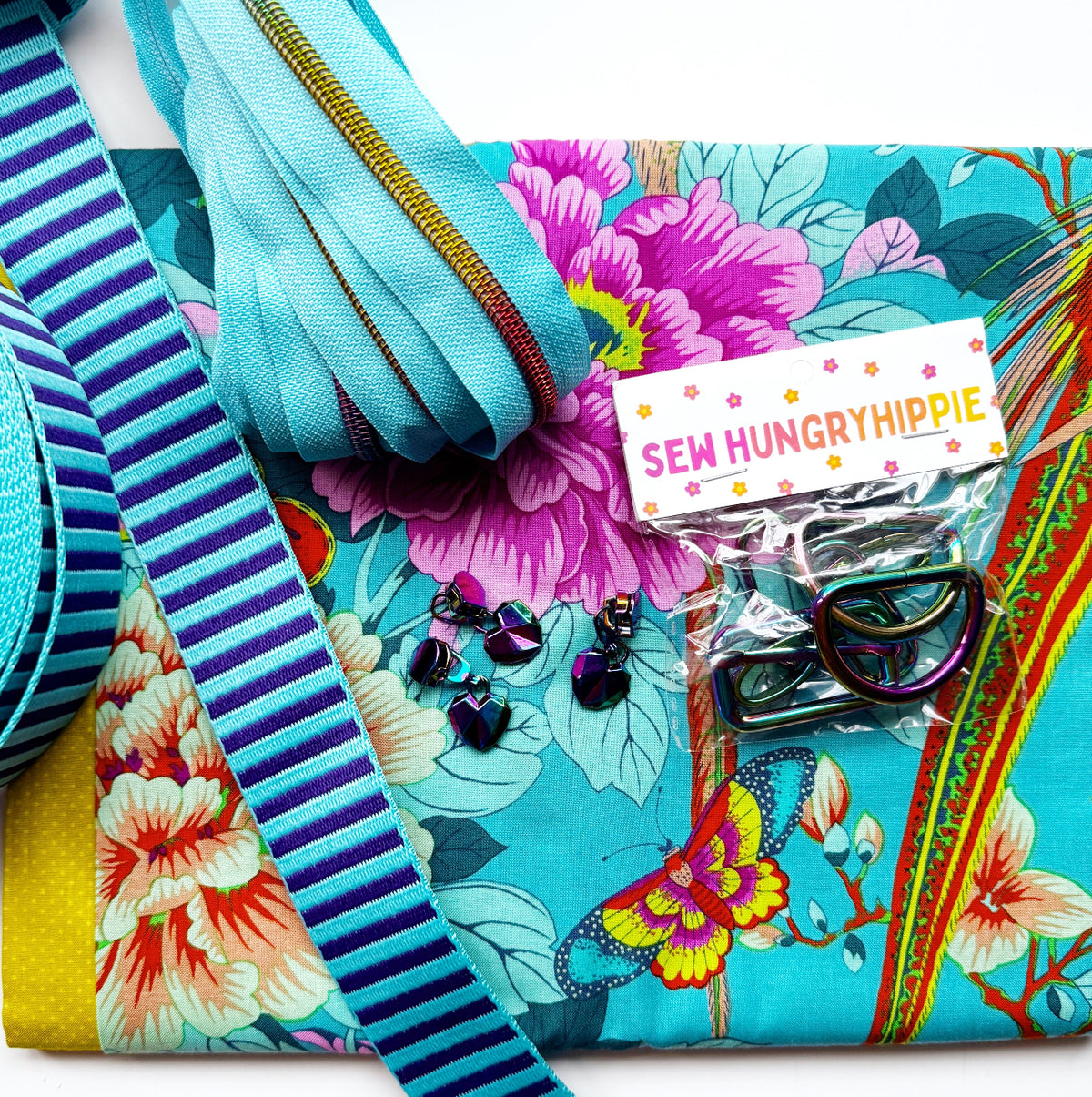 Sew Hungry Hippie bag-making supplies arranged on turquoise floral fabric featuring pink and peach peonies, green leaves, and a colorful butterfly. Includes purple and turquoise striped webbing, turquoise zipper tape with rainbow teeth, rainbow heart-shaped zipper pulls, and iridescent rainbow metal D-rings and sliders. 