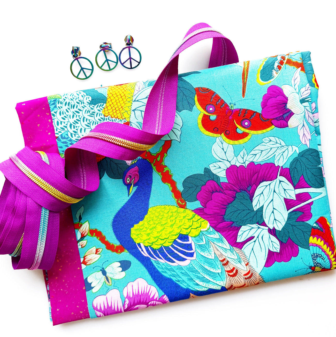 Turquoise cotton fabric featuring colorful peacocks and tropical flowers paired with fuchsia zipper tape, coordinating fabric, and iridescent peace sign hardware for bag making and sewing projects.