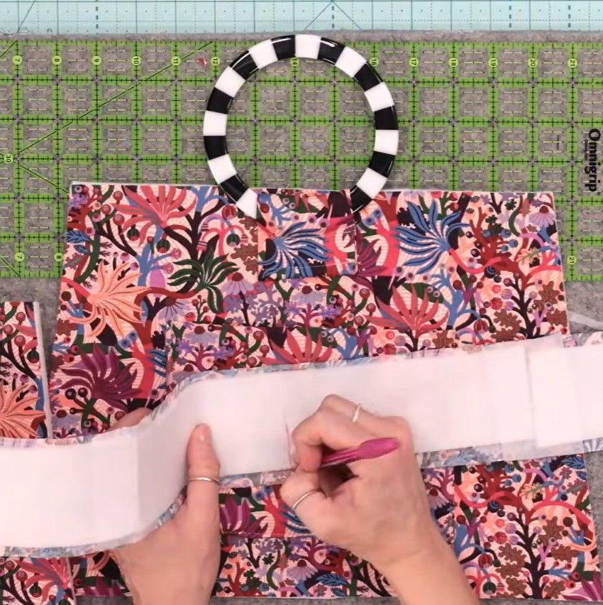 A person's hands are shown folding a white paper template over a colorful floral patterned fabric, with a black and white striped circular ring and a green gridded cutting mat in the background.