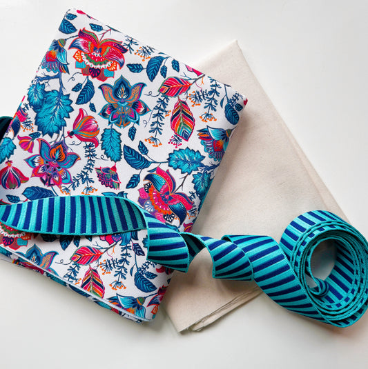 A folded white fabric item with a colorful floral and bird print, paired with a rolled teal and blue striped ribbon, placed on a light beige cloth against a white background.