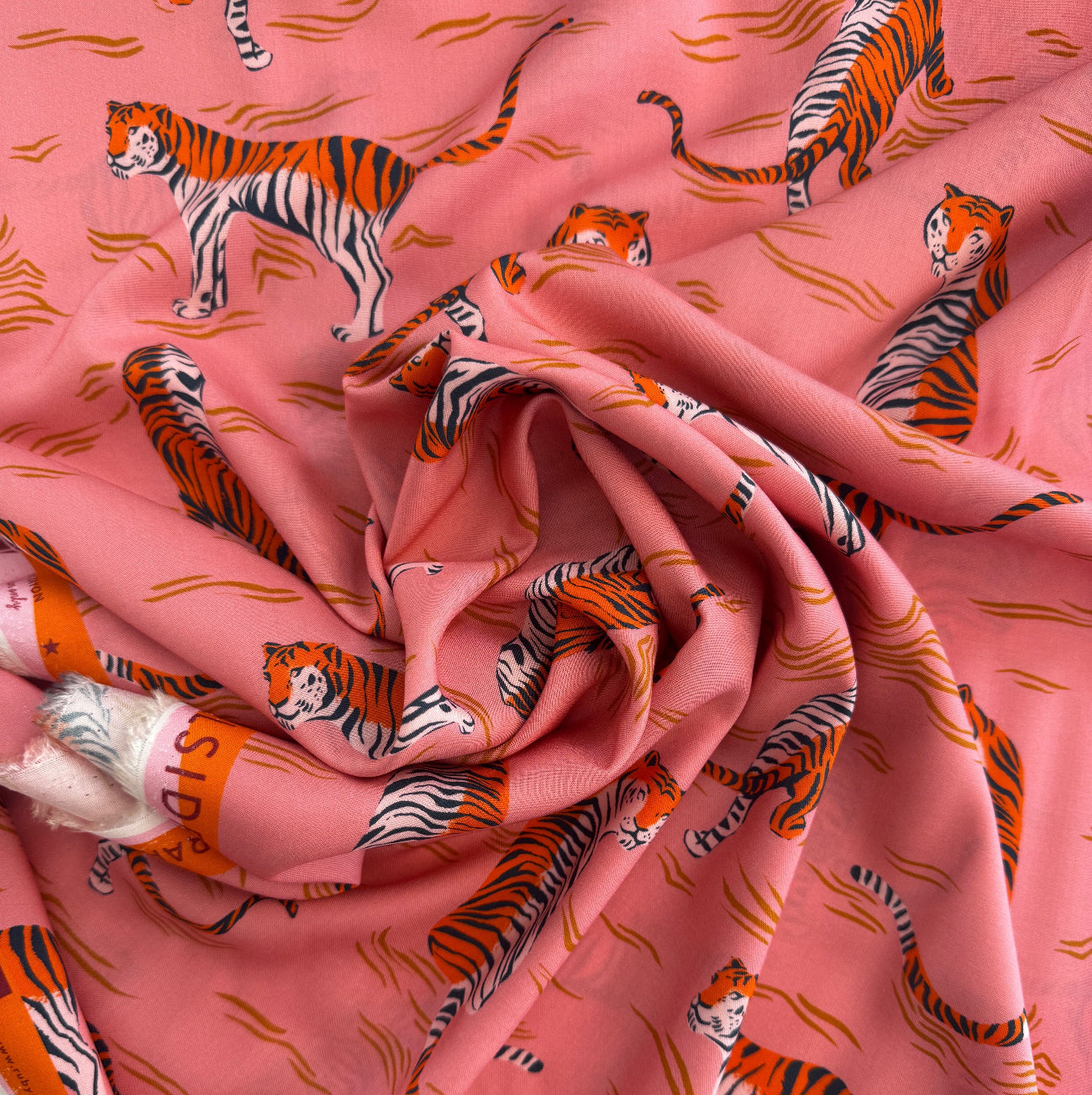 A high-angle, close-up shot of soft pink rayon fabric patterned with stylized orange-and-black tigers and wavy horizontal lines.