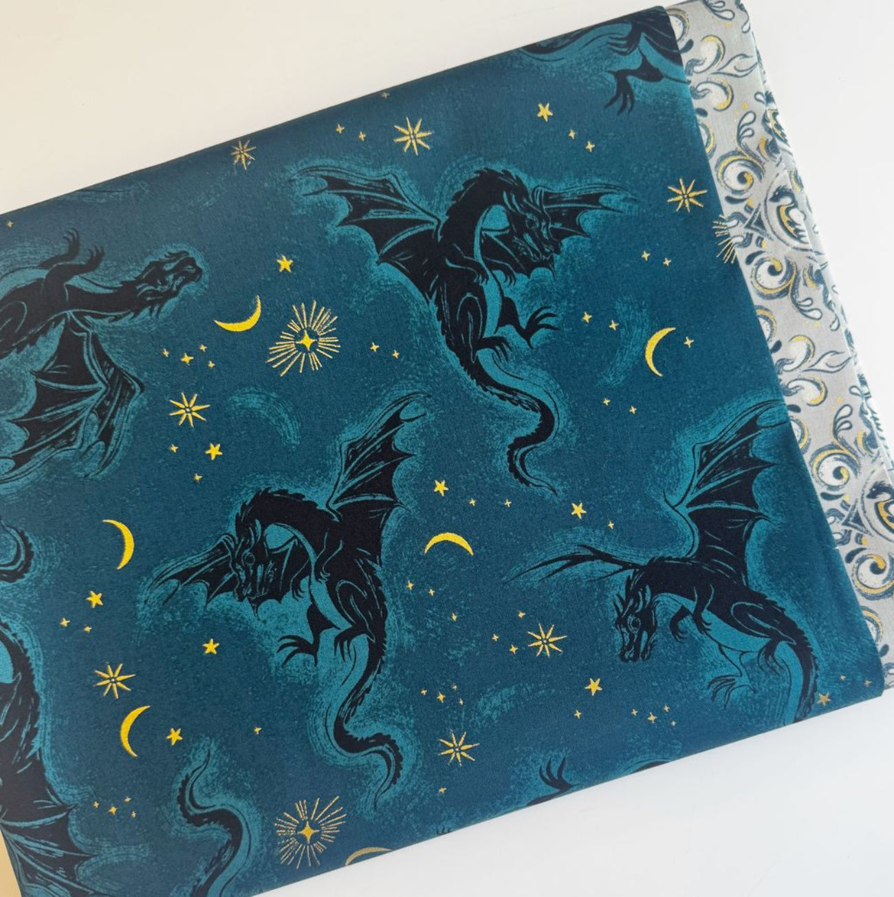 A 2-yard fabric bundle featuring Ruby Star Society's Fire Glow collection. The main fabric shows the "Dragon Flight" print in a deep teal colorway, featuring dark silhouettes of flying dragons surrounded by metallic gold stars and crescent moons. Peeking out from underneath is a coordinating light grey and metallic gold ornamental paisley print fabric, perfect for quilting, garment sewing, or lining handmade bags.