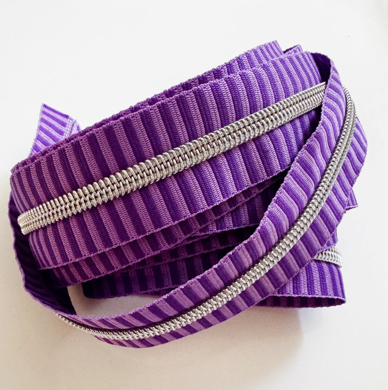 Number 5 continuous nylon coil zipper tape featuring a monochromatic dark and medium purple striped jacquard woven pattern with metallic silver teeth, loosely coiled against a white background.