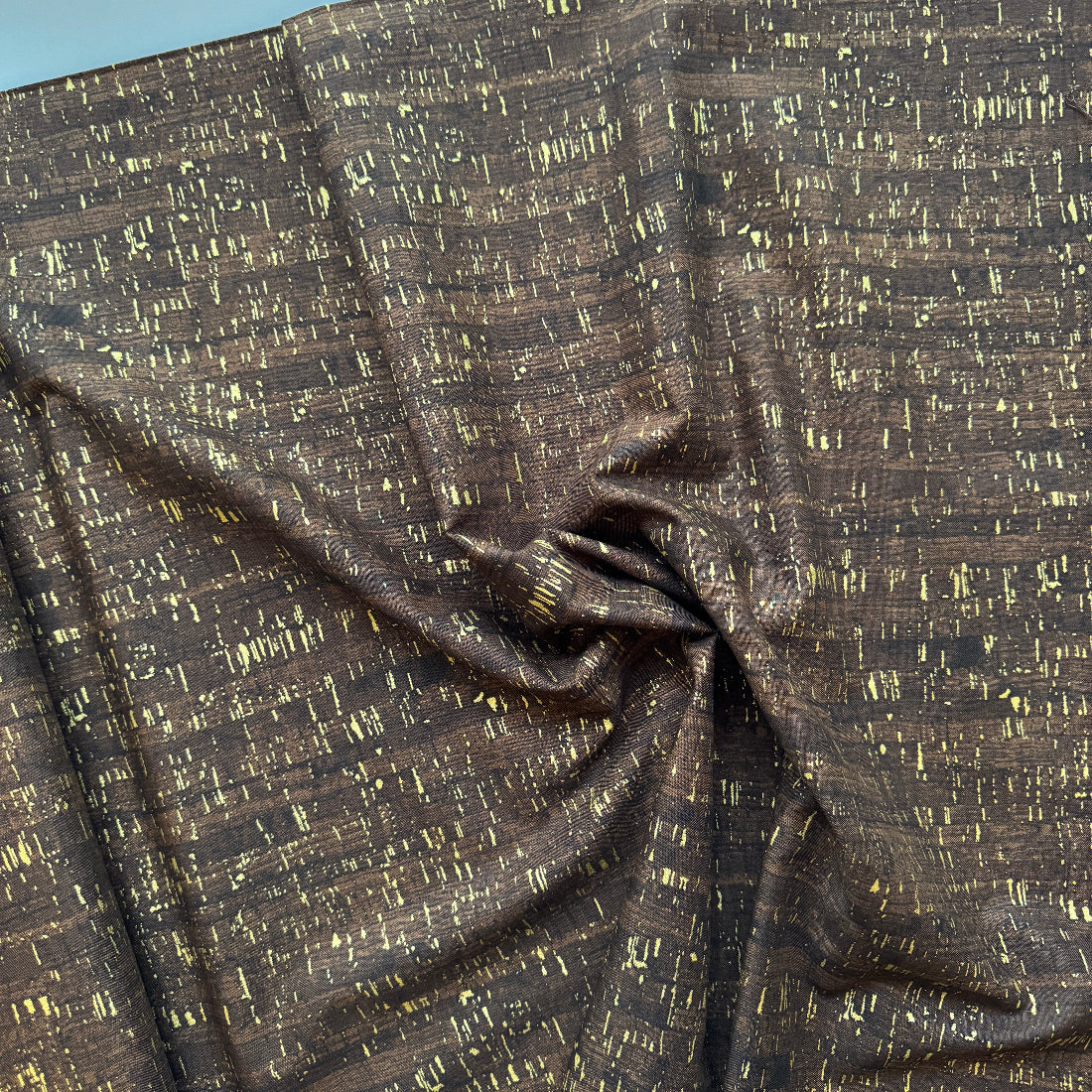 A close-up of draped fabric with a dark brown base and an all-over textured gold abstract pattern, gathered in the center to show its drape and sheen.