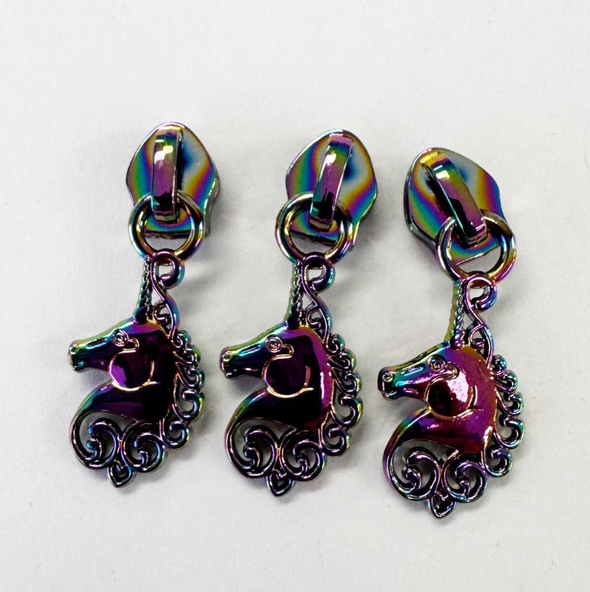 Three matching iridescent metallic unicorn-shaped zipper pulls or pendants arranged side by side on a plain white background.