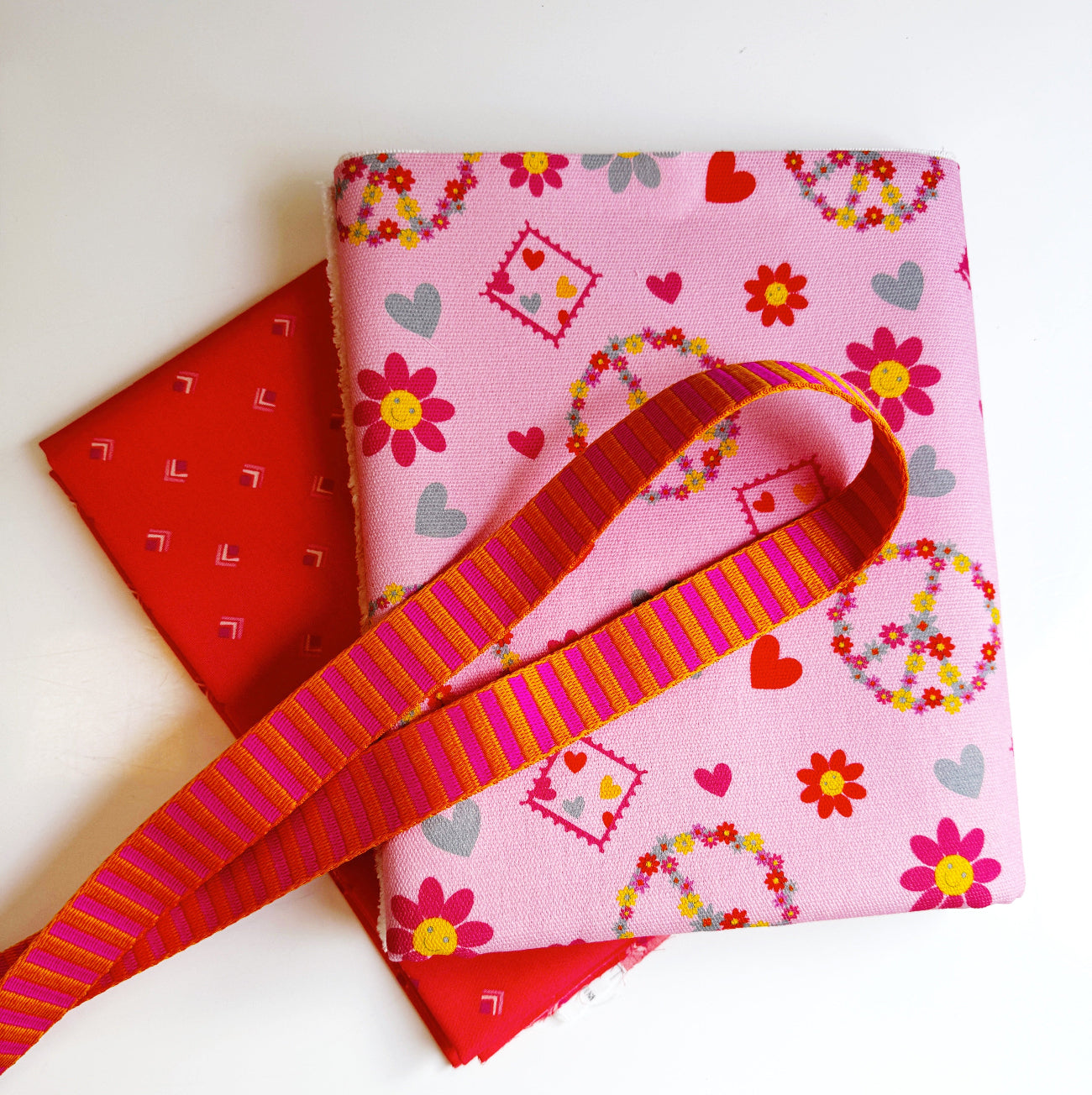 Peace heart print fabric with coordinating red fabric and pink-orange striped webbing for sewing a tote bag.
